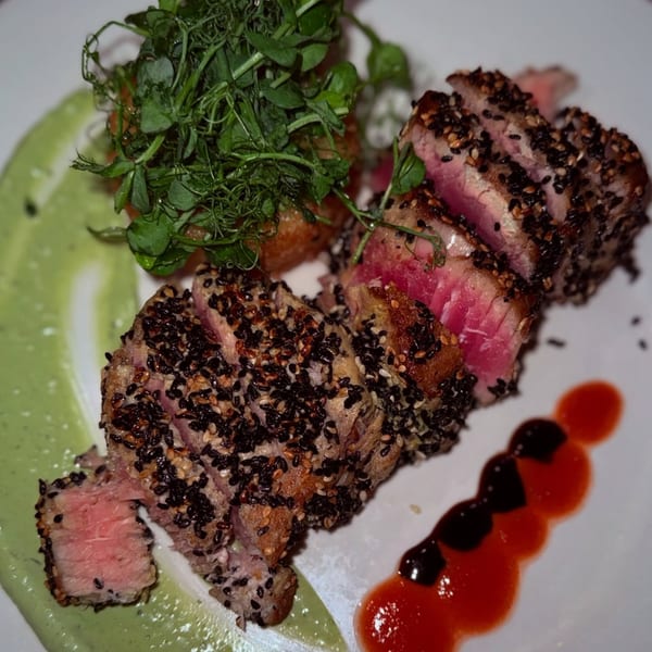 Seared tuna with sesame seeds, green puree, microgreens, and red and dark sauces on a white plate.