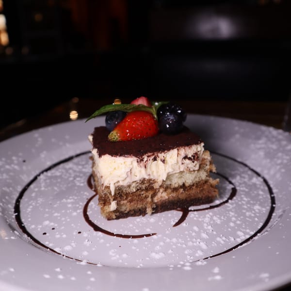 Tiramisu dessert with berries and mint on a white plate, drizzled with chocolate sauce.