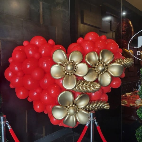 A large red heart-shaped balloon display with golden flowers and leaves, flanked by red velvet ropes.