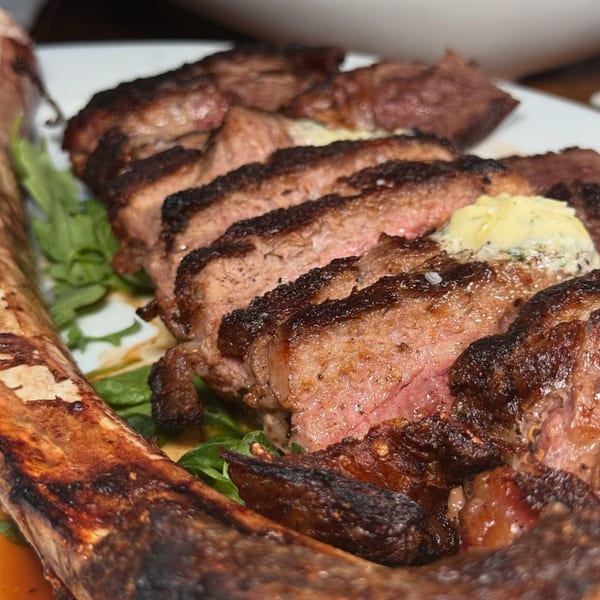 Sliced tomahawk steak with a bone, topped with melting butter and herbs, on a bed of greens.