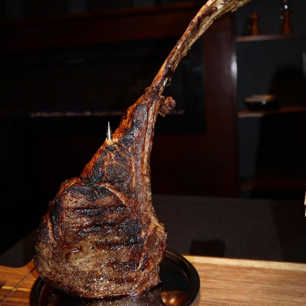 A large, perfectly cooked tomahawk steak with a long bone stands upright on a dark plate.