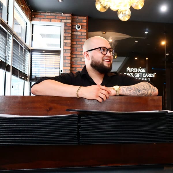 Smiling man with glasses and beard leaning on a wooden counter with menus at Jack's Bar And Steakhouse.